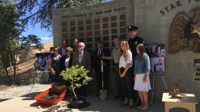 Family standing in front of memoral wall, small tree is being planted, Sheriff Alex Villanueva is holding a shoval.