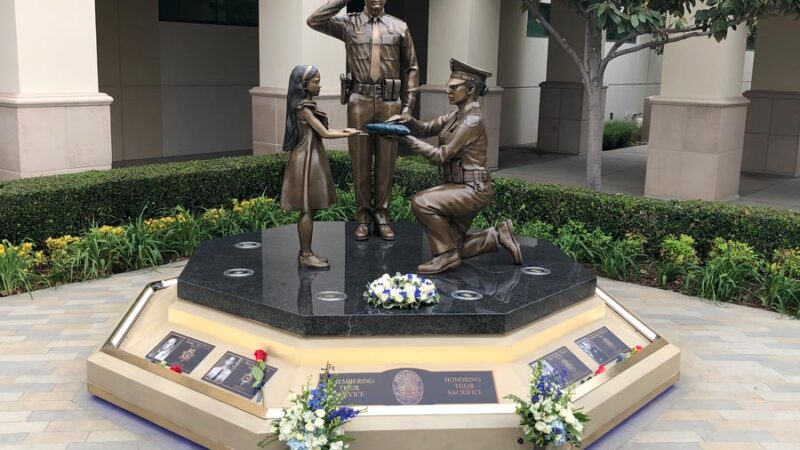 Image of the Buena Park Police departent memorial of fallen officers. Bronze statue of a saluting officer, another officer on one knee presenting a folded flag to a little girl. all cast in bronze.