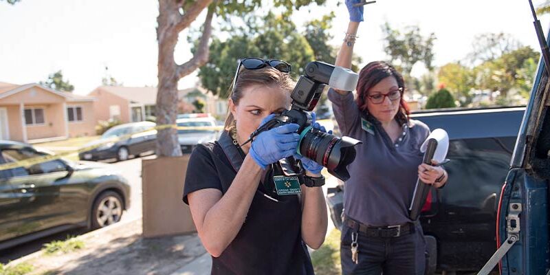 Picture of a woman in a black polo shirt with a Green Sheriff's Name plate preparing to take a picture with a camera, looking into the back of a truck that is just out of frame. The camera has a large lense and a flash. A second woman is holding up the back tail gate of the truck.
