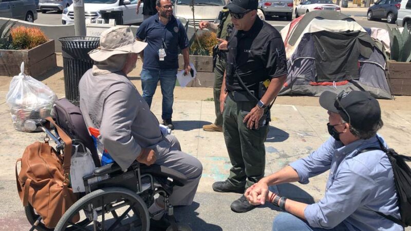 MET team deputy helping a homelss man in a wheelchair. there are three people from Vetran affairs talking to a man in a large white sun hat, he is dressed in a long sleeve gray shirt,the wheel chari has an orange backpack on the back, the homeless man is in his 60s.