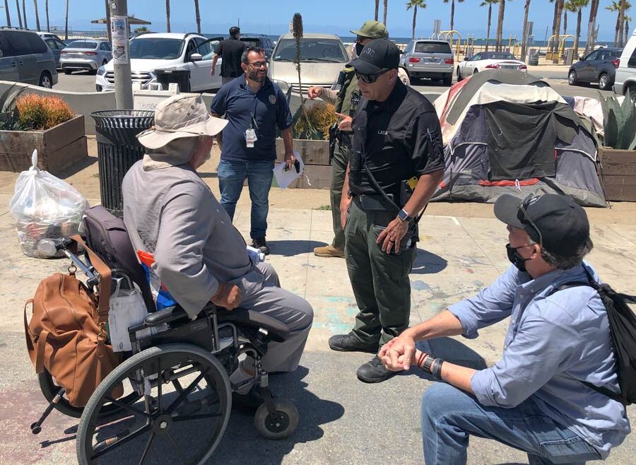 MET team deputy helping a homelss man in a wheelchair. there are three people from Vetran affairs talking to a man in a large white sun hat, he is dressed in a long sleeve gray shirt,the wheel chari has an orange backpack on the back, the homeless man is in his 60s.