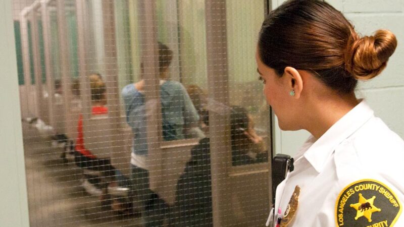 Visitation room has one stool and partitions on either side, facing a glass partition where inmates can visit with family. There is a Security Officer overlooking the row of visitation booths.