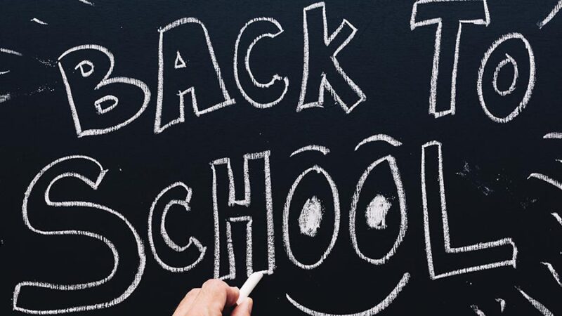 image of a hand writing on a chalk board, the board reads "BACK TO SCHOOL" and has a smile under the two o in the word "School".
