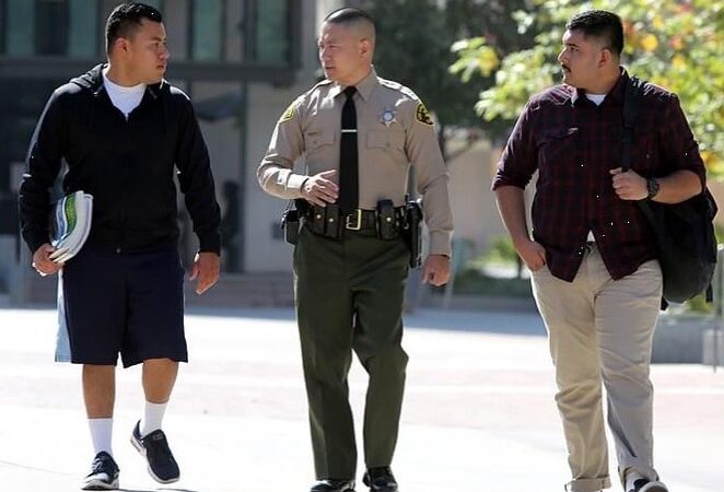 deputy sheriff is walking with two hispanic men down the street towards a camera. They are having a friendly conversation. The Deputy is wearing a tan longsleeve shirt with a black tie and green pants. there are two men on either side of the deputy. They are holding books and a backpack.