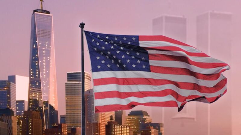 We Will Never Forget, September 11, 2001. Image of the american flag waving over the One Trade Center Tower on the left of flag and the Right side of flag are the Twin towers very faint over the orange purple sky.