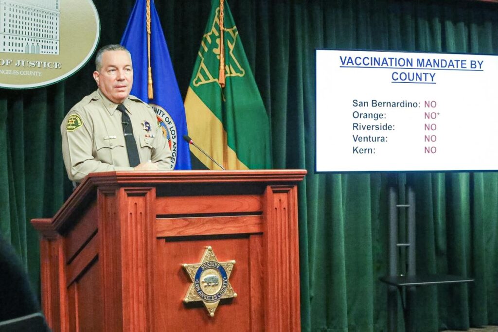Sheriff Alex Villanueva is standing behind a wood Podium speaking to media. He is standing infront of a green curtain next to County and California flags. There is a TV screen, on the screen the slide reads: "Vaccination Mandates by County: San Bernardino, no; Orange, no; Riverside, no; Ventura, no; Kern, no;'