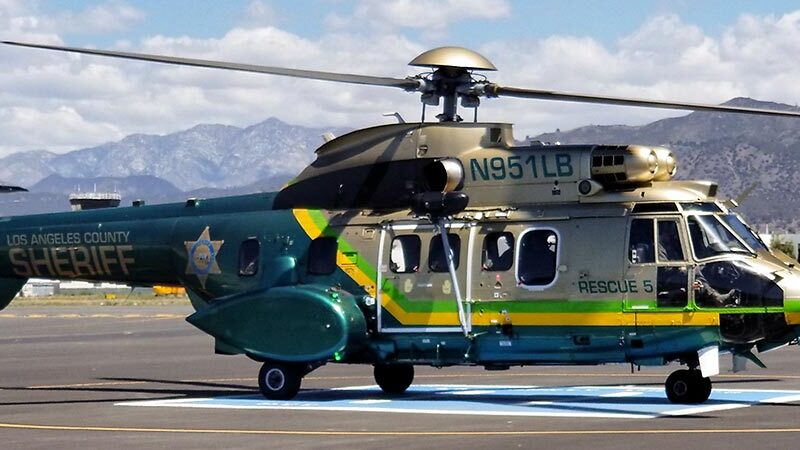 Image of Search and rescue heliocoptor on a runway. The heliocoptor is green and gold, is very large with a slideing door on the side where Search and Rescue personnel operate a wench. The markings on the heliocoptor are the Sheriff's star, and the words "Rescue 5".