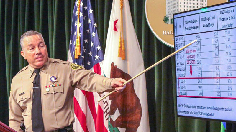 Sheriff Alex Villanueva in uniform pointing at a screen with a long wooden pointer. the screen has a budget in a table with numbers annotated in red. the text is too small to read from the picture taken.