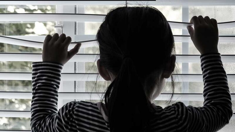 backlit picture of a child looking throgh a window with her hands on blinds, peering out.
