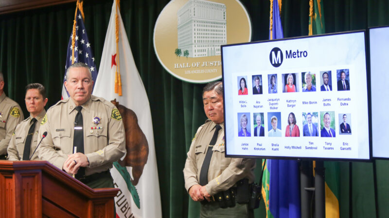 Sheriff is standing behind a podium. Undersheriff Murakami is over the sheriff's shoulder. A TV screen has an image of the METRO board of supervisors on it.