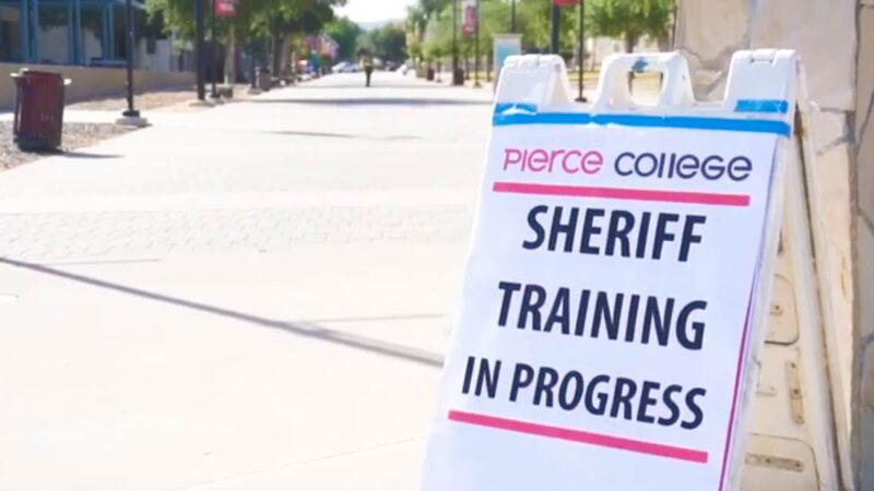Image of an A-frame sign with big black letters that read "Sheriff training in progress". The sign in the lower left side of the frame. The sign is infront of a college walk path.