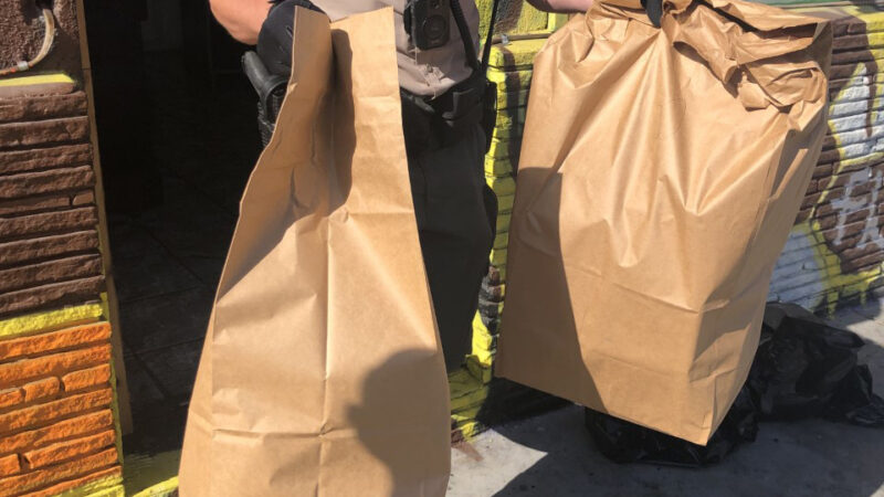 deputy is walking out of a business door holding two paper bags full of confiscated drugs.
