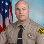 Captain Torres is dressed in a tan longsleeve shirt with a black tie. He is sitting infront of a blue background with the American flag visible behind his right shoulder. .