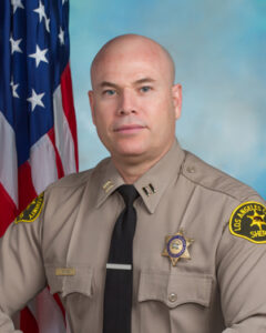 Captain Torres is dressed in a tan longsleeve shirt with a black tie. He is sitting infront of a blue background with the American flag visible behind his right shoulder. .