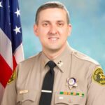 portrait of captain kehoe in uniform, tan shirt black tie. Sitting infront of a blue background.