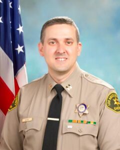 portrait of captain kehoe in uniform, tan shirt black tie. Sitting infront of a blue background.