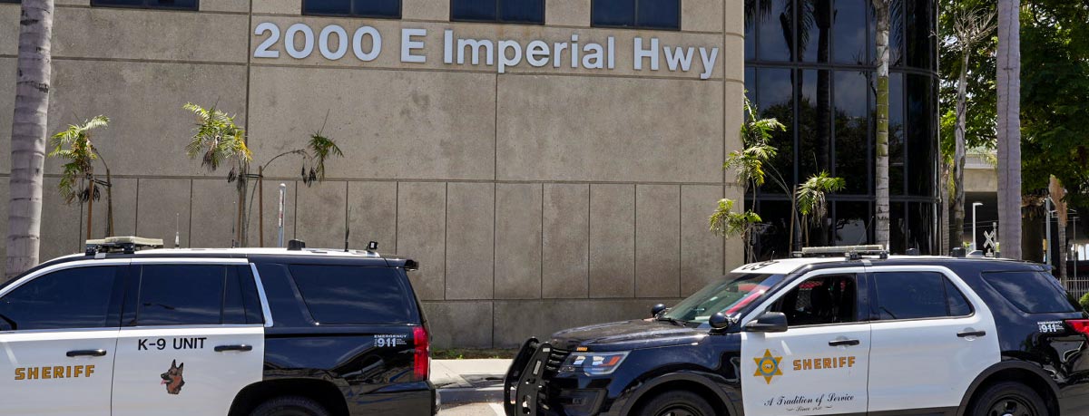 picture of two black and white Sheriff vehicles parked infront of the Transit Service's station. The building is tan marble with large address "2000 East Imperial Hwy."